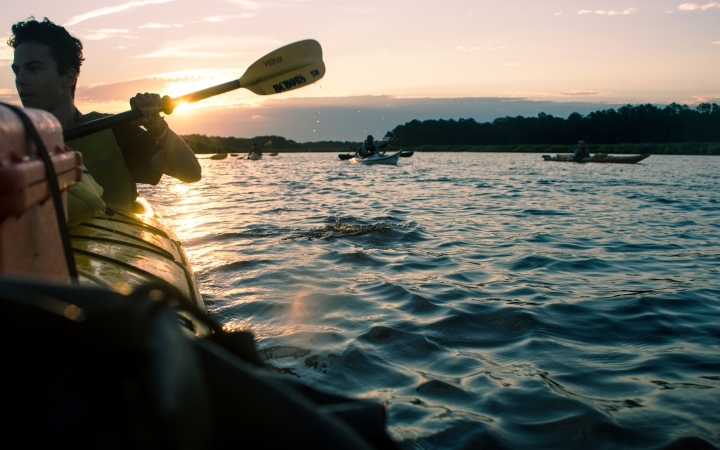 in the foreground, a person paddles a canoe ahead of several others while the sun sets behind them
